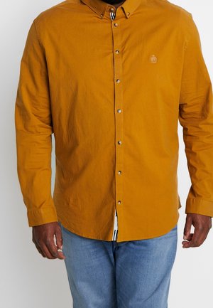 Mosterdgele blouse met lange mouwen en een kraag, voorzien van drukknopen en een klein logo op de borst. Denim blauwe jeans.