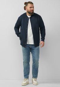 Marine Bomberjacke mit Druckknöpfen, getragen über einem weißen T-Shirt, kombiniert mit hellblauen Jeans und beigen Sneakers. Glatte Textur, schlichtes Design.