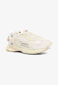 Crème-kleurige Lacoste sneakers met een gestructureerd ontwerp, voorzien van lichtgele accenten en bedrukte tekst. De zool heeft een gepatternde profiel.