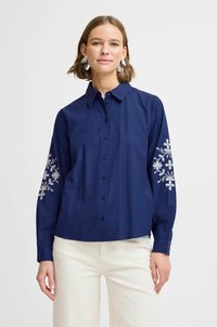 Navy blue button-up shirt with long sleeves featuring white floral embroidery on the sleeves. Made of smooth fabric, relaxed fit design.