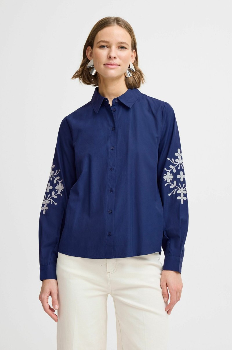 Camisa de botón azul marino con mangas largas que presenta bordado floral blanco en las mangas. Fabricada con tela suave, diseño de corte relajado.