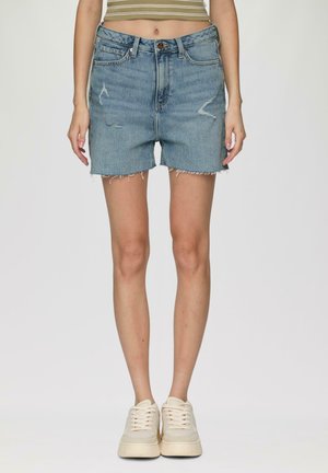 Shorts en denim clair avec taille haute, ourlet effiloché et détails usés à l'avant. Portés avec des baskets de couleur crème.