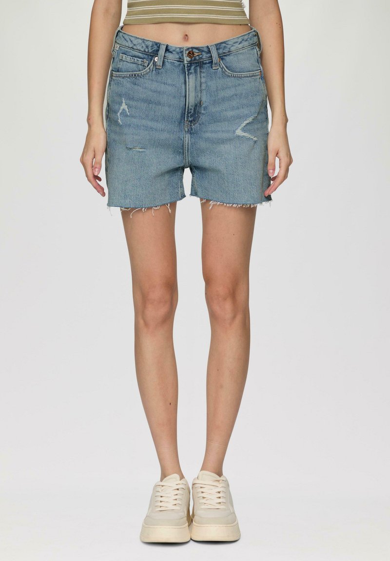Jasnoniebieskie szorty dżinsowe z wysokim stanem, postrzępionym wykończeniem i przetartymi detalami z przodu. Noszone z kremowymi trampkami.