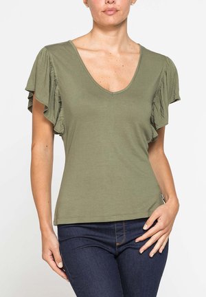 Donna che indossa un top verde oliva a scollo a V con maniche corte arricciate e jeans aderenti blu scuro su uno sfondo semplice.