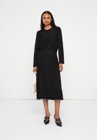 Schwarzes Maxikleid mit langen Ärmeln und strukturiertem Muster, bis zur Mitte der Wade reichend, mit Fransenverzierung. Kombiniert mit schwarzen Schlappen und einer kleinen rosa Handtasche.