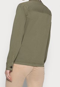 Osoba v olivově zeleném triku s dlouhým rukávem a béžových kalhotách, zobrazená zezadu na jednobarevném bílém pozadí.