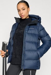 Mørkeblå dynejakke med hætte, med quiltet design, lynlås foran og logoaccent. Båret over en mørk strikket sweater.