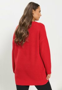 Pull en tricot rouge à coupe décontractée, avec un motif texturé, des épaules tombantes et des poignets et ourlets côtelés. Stylisé avec un pantalon noir.