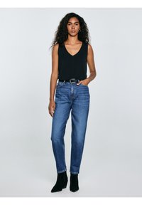 Vrouw met krullend haar, gekleed in een zwart mouwloos topje, een high-waisted blauwe spijkerbroek, een zwarte riem en zwarte enkellaarsjes, staand met één hand in haar zak.