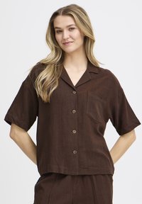 Chemise marron à manches courtes en tissu léger, avec un col, une poche poitrine et des boutons marron sur le devant.