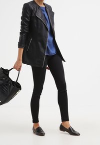 Veste en cuir noir avec fermeture éclair asymétrique, haut en tricot bleu, jeans skinny noirs et mocassins noirs à talons pointus, tenant un grand sac fourre-tout noir.