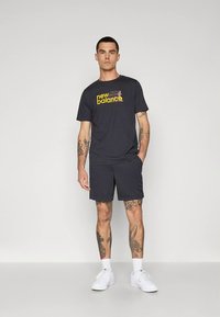 Mörkgrå New Balance t-shirt för män med gul logotyp, matchad med motsvarande shorts. Bomullstyg, korta ärmar, ledig passform och vita sneakers.