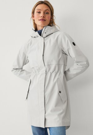 Vrouw draagt een lichtgrijze regenjas met capuchon, voorflapzakken en elastische taille, staande tegen een egale witte achtergrond.