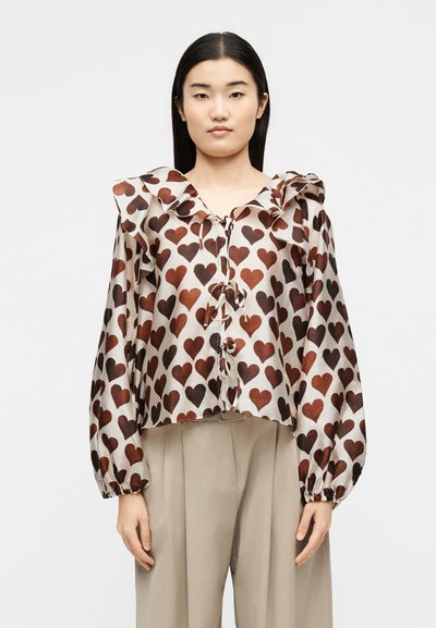 Γυναίκα με ίσια μαύρα μαλλιά φοράει μπεζ πουκάμισο με καφέ σχέδια καρδιών, γιακά με βολάν και μπεζ παντελόνι με ψηλή μέση και φαρδιά πόδια.
