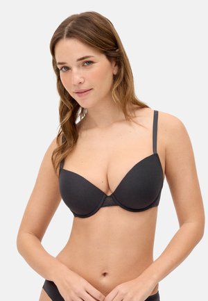 Mujer joven con cabello castaño claro, vistiendo un sujetador negro con copas moldeadas, posando frente a un fondo neutro y claro.