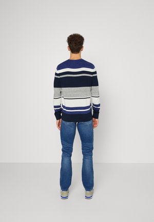Homme debout, dos tourné, portant un pull rayé à manches longues, un jean bleu et des baskets grises sur un fond uni clair.