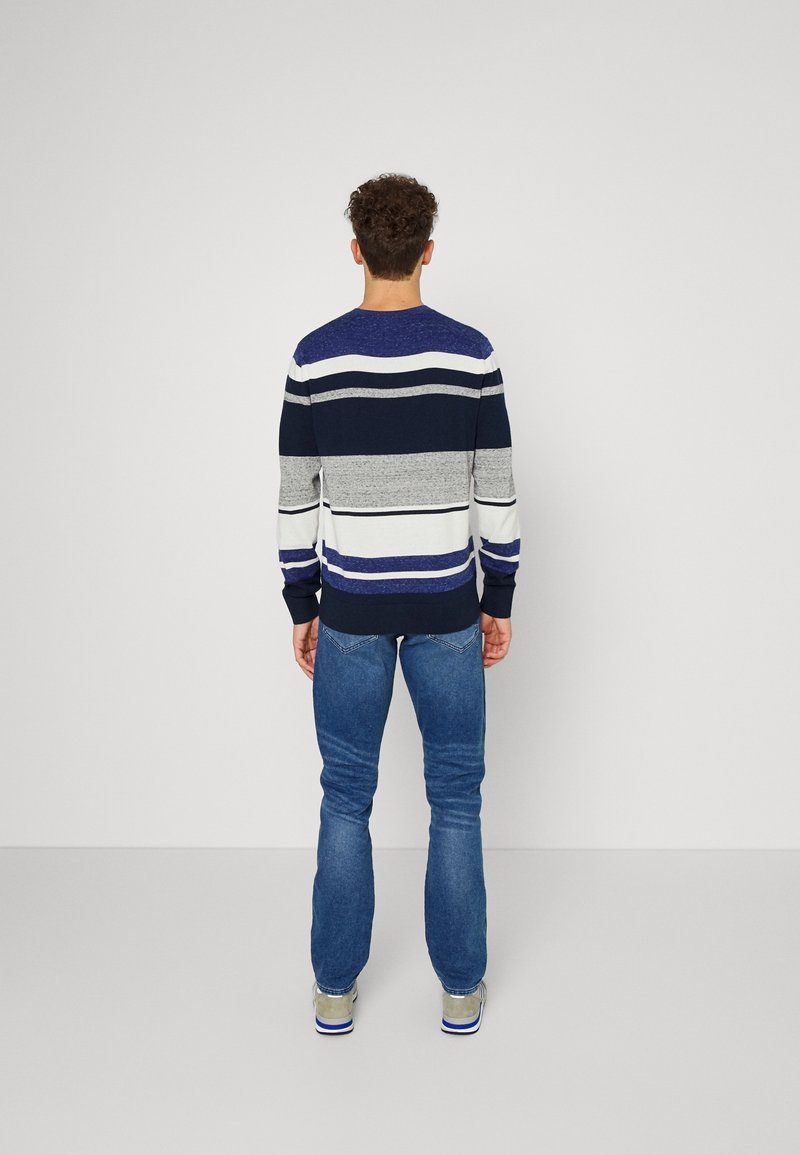 Homme debout, dos tourné, portant un pull rayé à manches longues, un jean bleu et des baskets grises sur un fond uni clair.