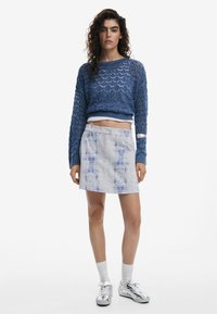 Stickad blå tröja med geometriska mönster, tillsammans med en kort ljusblå kjol med en färgliknande textur, och silvriga sneakers.