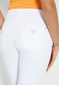 Jean en denim blanc avec coutures décoratives sur les poches, arborant une étiquette de logo de marque triangulaire. Texture lisse et coupe ajustée.