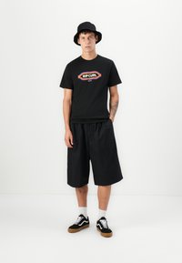 Svart bomulls t-skjorte med "RIP CURL" logo i fargerik skrift, sammen med løse svarte shorts og svarte joggesko med hvite detaljer.