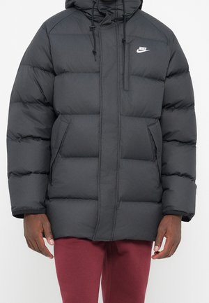 Giacca imbottita nera Nike con cappuccio e coulisse indossata da una persona con pantaloni color bordeaux, che mostra la cerniera frontale e le tasche laterali.