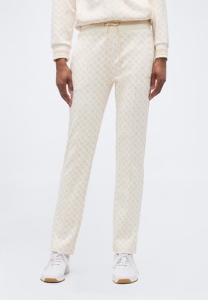 Henkilö, joka käyttää kermaisen värisiä housuja, joissa on hienovarainen kuvio, vastaa vyötärönyöri ja valkoiset tennarit, seisoo yksinkertaisen taustan edessä.