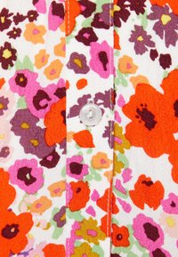 Bílá látka s oranžovým, růžovým, fialovým a žlutým květinovým vzorem, s centrálním bílým knoflíkem a prošitými švy.