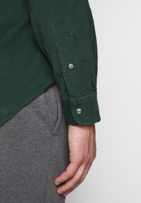 Zelená kordurová košile s knoflíkovou manžetou, která má jemnou ribkovou texturu a zakřivený spodní lem, v kombinaci se šedými tepláky.