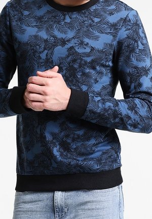 Mand iført blå sweatshirt med sort blomster- og bladmønster, sort krave og manchetter samt lyseblå jeans, hænderne foldet foran.