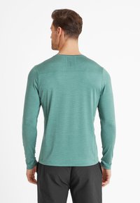 Man wearing teal long-sleeve shirt and black pants, standing facing away against a plain white background.