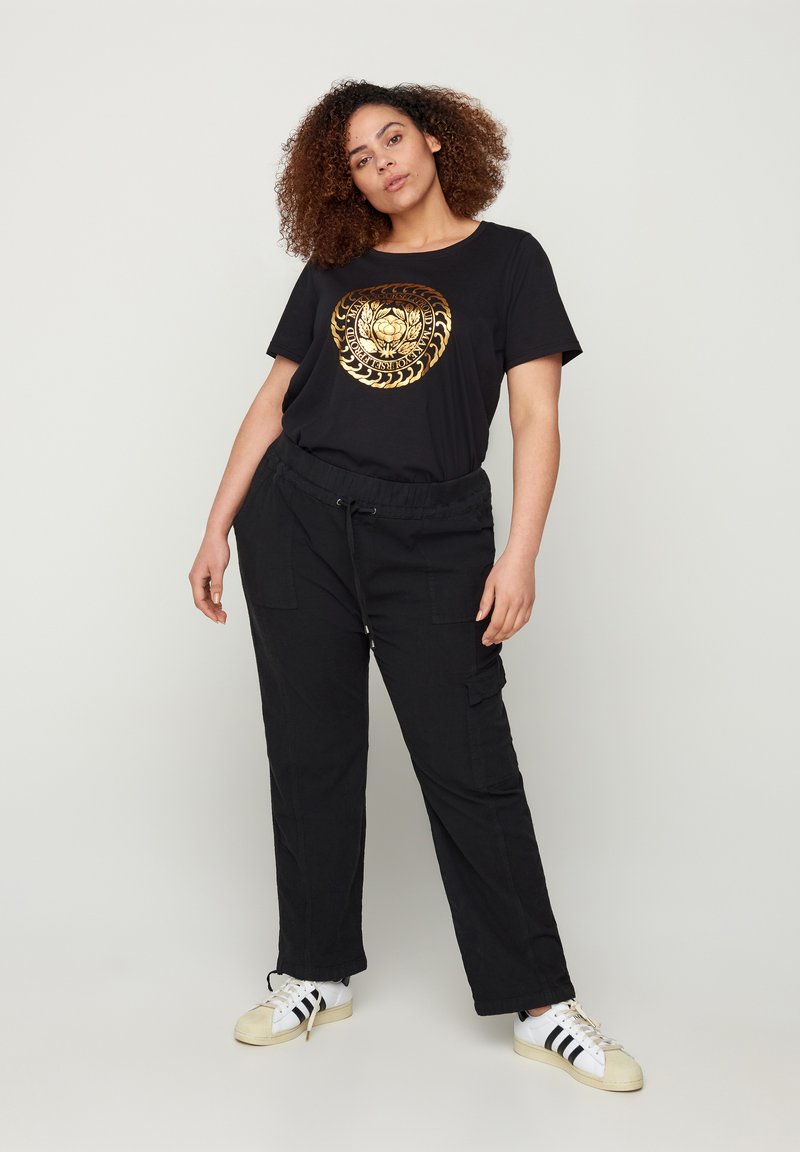 T-shirt nera con stampa grafica dorata, abbinata a pantaloni cargo neri e sneaker bianche con strisce nere. Vestibilità relax; stile casual.