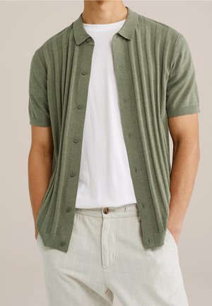 Mann trägt eine kurzärmelige olivgrüne Strickjacke mit Knöpfen über einem weißen T-Shirt und hellbeigen gestreiften Hosen, steht mit Händen in den Taschen.