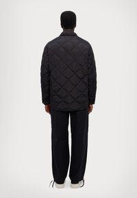 Giacca nera imbottita con motivo a rombi, materiale leggero, design del colletto e vestibilità comoda, abbinata a pantaloni scuri e scarpe chiare.