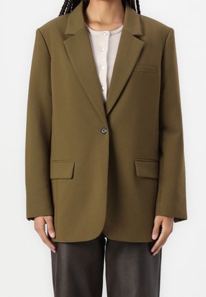 Personne portant un blazer vert olive à un bouton sur un haut beige clair boutonné et un pantalon marron foncé, debout devant un fond blanc.