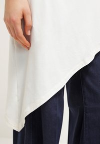 White asymmetrical top made of soft fabric, featuring a rounded hem. Close to navy-blue pants visible at the bottom. Hand lightly touches the fabric.