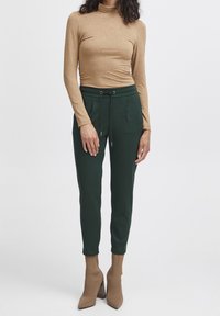 Koffiebruin langarmtop met een aansluitend ontwerp, gecombineerd met donkergroene joggingbroeken met een trekkoord en twee voorkruiszakken. Accessoires omvatten tan hoge hakken.
