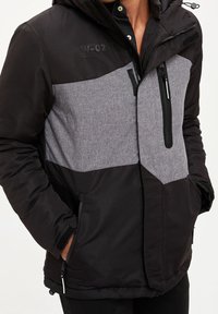 Černá a šedá zateplená bunda s kapucí, s předním zipem, dvěma bočními kapsami a kontrastním texturovaným panelem.
