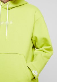 Kapotová mikina v limetkově zelené barvě vyrobená z bavlny, s přední kapsou, stahovacími šňůrkami a textem "just do it." Logo Nike na manžetě.
