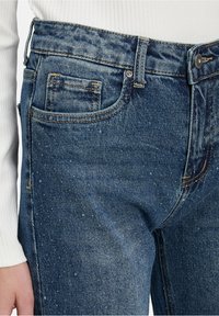 Jean en denim taille haute dans un lavage bleu foncé, avec une poche avant, des coutures contrastantes et une fermeture à boutons.