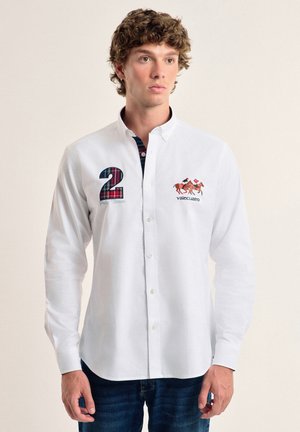 Camisa blanca de botones con mangas largas, que presenta un número "2" de cuadros y una ilustración de un caballo en el pecho. Material de algodón suave.