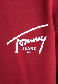 Felpa rossa con logo bianco che reca la scritta "Tommy Jeans" e un piccolo design a bandiera. Il tessuto sembra morbido e ha una texture liscia.