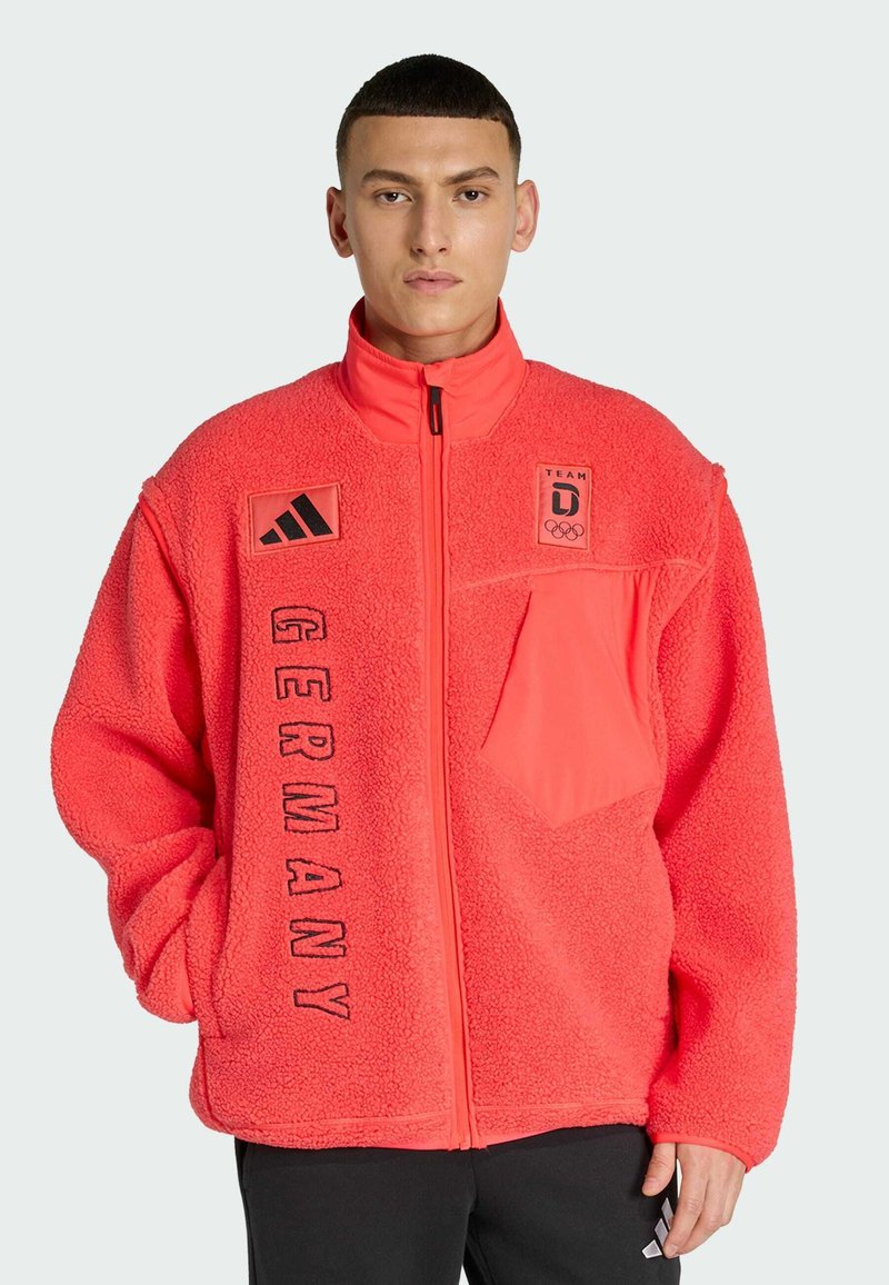 Helle Korallen-Fleecejacke mit hohem Kragen, Reißverschluss, Fronttasche und gesticktem "GERMANY"-Schriftzug. Enthält Adidas-Logos.