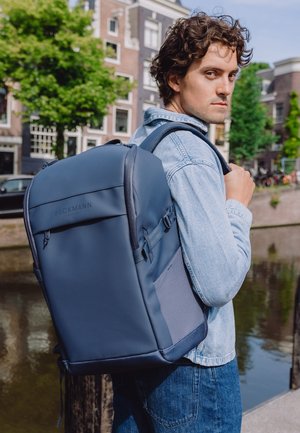 Ung mann i denimjakke som bærer en stor blå Beckmann-sekk nær en kanal i urbant område med trær og bygninger.