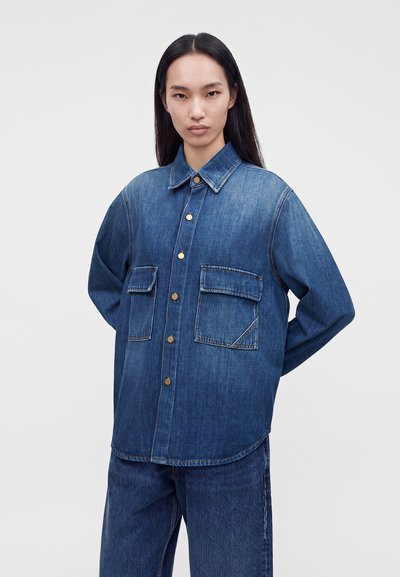 Femme aux longs cheveux foncés portant une veste en jean bleue oversize avec de grandes poches devant et un pantalon en jean assorti sur un fond uni.