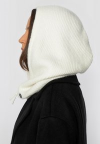 Witte geribbelde gebreide kap met trekkoorddetail, gedragen over een zwarte jas, met een gladde textuur en een nauwsluitende pasvorm rond het gezicht.