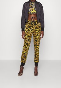 Svart bomberjacka över en kortärmad tröja med svart och guld grafik, kombinerad med svarta byxor med ett gult barockmönster och svarta muddar.