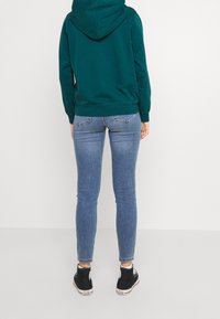 Zielona bluza z kapturem, jasnoniebieskie wąskie dżinsy z tylnymi kieszeniami oraz czarne sneakersy z białymi akcentami. Widok z tyłu.