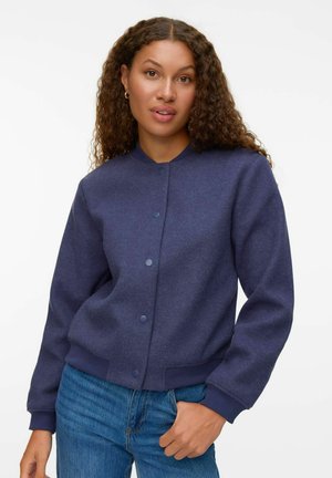 Veste bomber bleu marine texturée avec un col rond, fermeture à boutons et poignets côtelés, associée à un jean bleu clair.