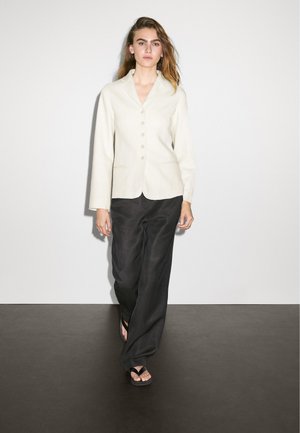 Woman walking in white buttoned blazer, loose black pants, and black sandals against a plain white wall and dark floor.