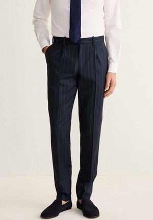 Hombre con pantalones azul marino de rayas finas, camisa blanca, corbata azul oscura y mocasines negros, de pie con las manos en los bolsillos.